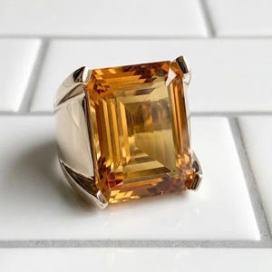 May include: A silver ring with a large, rectangular-cut citrine gemstone. The gemstone is a vibrant golden-orange colour. The ring is set with four prongs. The ring is displayed on a white surface.