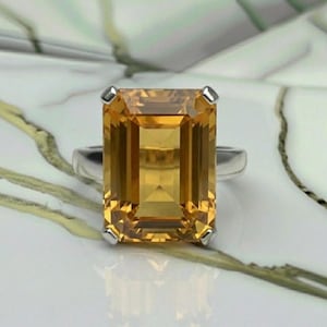 May include: A silver ring featuring a large, rectangular-cut, golden-yellow gemstone. The gemstone is set with four corner prongs, and the band is a simple, polished silver. The ring is displayed on a white and gray marbled surface.