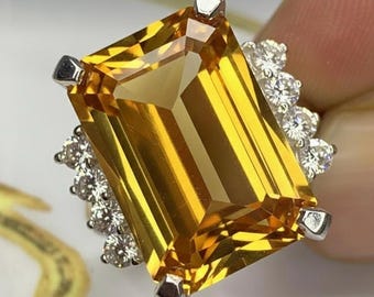 May include: A large, rectangular-cut citrine gemstone ring with a vibrant yellow hue. The ring features a halo of small, round, clear gemstones and silver-colored prongs. The ring is a statement piece of jewelry.