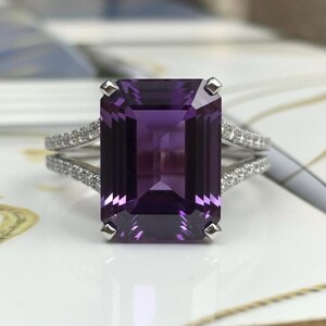 May include: A silver ring featuring a large, rectangular, deep purple gemstone. The band splits into two parallel rows, each encrusted with small, clear gemstones. The ring is set against a soft, blurred background.