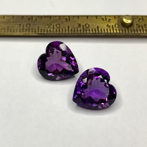 May include: Two heart-shaped amethyst gemstones, one on top of the other, on a white surface.