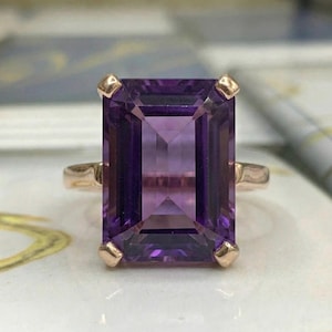 May include: A rose gold ring featuring a large, rectangular, faceted amethyst gemstone. The amethyst is a deep purple color with a lighter center. The ring has a simple band and four prongs at each corner of the gemstone.