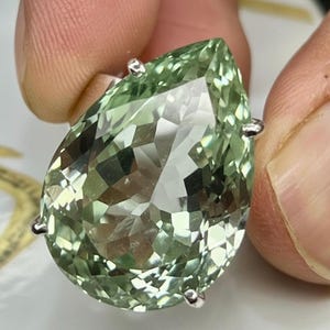 Può includere: Un anello con una pietra preziosa verde chiaro sfaccettata a forma di pera. La pietra preziosa è tenuta in posizione da quattro griffe d'argento. L'anello è tenuto tra due dita. La pietra preziosa ha un alto grado di chiarezza e brillantezza.