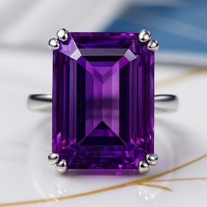 May include: A striking ring featuring a rectangular, deep purple amethyst gemstone. The gemstone is set in a silver band, with small, round silver accents at each corner. The ring is displayed on a white surface with gold detailing.