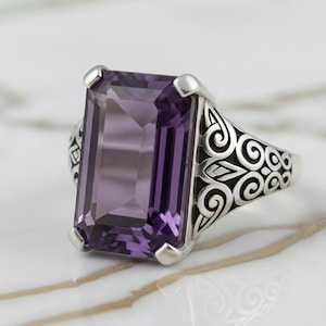 Puede incluir: Un anillo de plata con una gran gema púrpura rectangular facetada. El anillo tiene un diseño ornamental y arremolinado en la banda y alrededor del engaste de la gema. La gema es de un tono púrpura intenso.