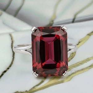 May include: A large, rectangular-cut, deep red gemstone ring with a silver band. The gemstone is set with four prongs. The ring is displayed on a white and gold marbled surface. The gemstone's facets reflect light.