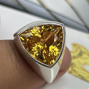 Pode incluir: Um anel de prata com uma grande pedra preciosa amarela triangular facetada. A pedra preciosa está engastada numa banda de prata polida, mostrando o seu corte brilhante e a sua cor quente. O design do anel é moderno e atraente.