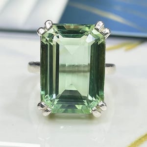 Op de afbeelding: Een zilveren ring met een grote, rechthoekige, lichtgroene edelsteen. De edelsteen is facetgeslepen, met een heldere, smaragdgroene uitstraling. De zetting van de ring heeft decoratieve zilveren accenten op elke hoek van de edelsteen.