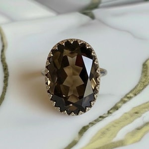 Peut inclure: Une bague ovale ornée d'une grande pierre de quartz fumé facettée. La pierre précieuse, de couleur marron foncé, est sertie dans un sertissage métallique décoratif et texturé. La bague est posée sur une surface en marbre blanc et or.