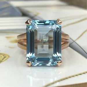 Pode incluir: Um anel de ouro rosa com uma grande pedra preciosa retangular azul clara. A pedra preciosa tem múltiplas facetas, refletindo a luz. A banda do anel divide-se em duas bandas paralelas em cada lado da pedra preciosa.