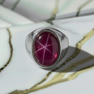 May include: A silver ring with a large, oval-shaped, deep red gemstone. The gemstone features a six-rayed star pattern. The ring has a thick band and a polished finish, showcasing a classic and elegant design.