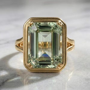 Puede incluir: Un anillo de oro con una gran piedra preciosa de color verde claro, de corte esmeralda rectangular, engastada en un bisel con cuatro garras en las esquinas. La banda tiene un diseño dividido que se conecta a los lados del engaste rectangular.
