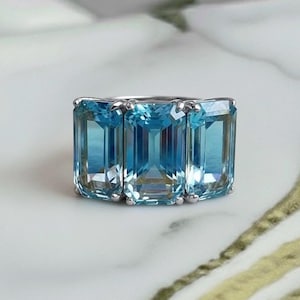 May include: A silver ring with three rectangular, light blue gemstones. The gemstones are emerald-cut and have a clear appearance. The ring is displayed on a white and gold marble surface. The gemstones are a light shade of blue.