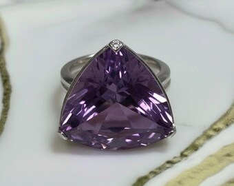 May include: A silver ring featuring a large, triangular, faceted amethyst gemstone. A small diamond is set at the top of the amethyst. The ring is set against a white and gold marble surface.