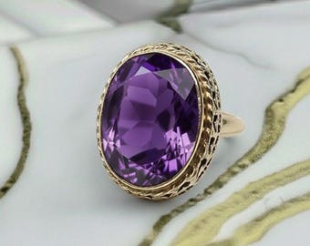 Puede incluir: Un anillo de oro con una gran piedra preciosa púrpura de talla ovalada. La piedra preciosa está engastada en un diseño de filigrana, con una banda de oro. El anillo se exhibe sobre una superficie de mármol blanco y dorado.