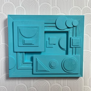 May include: A teal blue framed artwork with a layered geometric design. The artwork features circles, squares, and triangles in various sizes and positions.