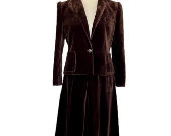 May include: A vintage brown velvet suit, consisting of a blazer and a knee-length skirt, displayed on a mannequin. The blazer has a single button closure and two front pockets. The skirt is pleated. A cream-colored turtleneck is visible beneath the blazer.