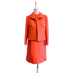 Op de afbeelding: Een feloranje tweedelig pak bestaande uit een kort jasje en een bijpassende rok. Het jasje heeft een kraag, lange mouwen en twee klepzakken aan de voorzijde. De rok valt tot op de knie en is voorzien van verticale naaddetails.