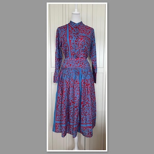 Puede incluir: Un vestido de manga larga con estampado azul y rojo y falda fruncida. El vestido tiene cuello y cierre de botones.