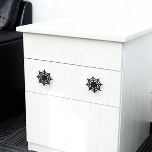 Może przedstawiać: Biała komoda z trzema szufladami i czarnymi uchwytami w kształcie pajęczyny. Komoda stoi na tle białej ściany, z czarną skórzaną sofą i rośliną doniczkową w tle.