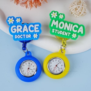 May include: Two retractable badge reels with a blue and yellow watch. The blue reel has a blue and white name tag with the text "GRACE DOCTOR *" and the yellow reel has a green name tag with the text "MONICA STUDENT *"