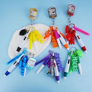 Könnte beinhalten: Eine Sammlung von einziehbaren Ausweisrollen mit verschiedenen Anhängern und Schlüsselanhängern. Zu den Anhängern gehören mit Glitzer gefüllte Infusionsbeutel, Namensschilder und Wortanhänger. Stifte mit farbigen Akzenten sind an den Schlüsselanhängern befestigt.