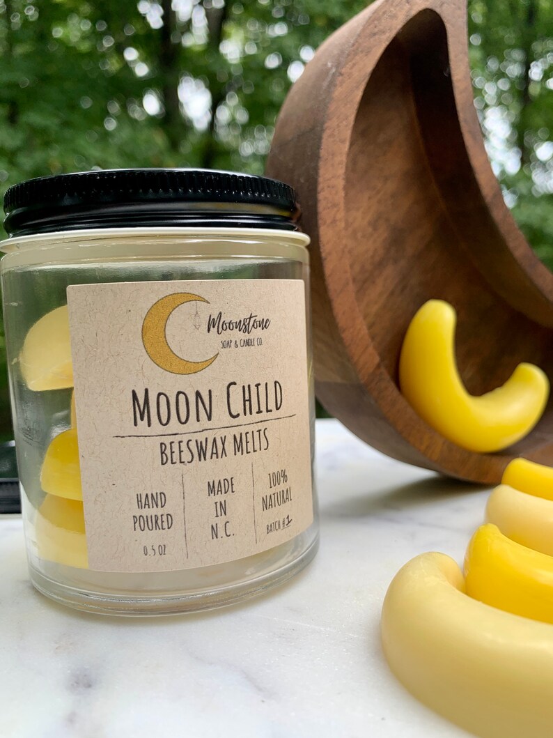 May include: A glass jar filled with yellow beeswax melts labeled "Moon Child Beeswax Melts". The label also states "Hand Poured", "Made in N.C.", "100% Natural", and "Batch #1".