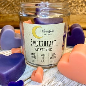 May include: A glass jar filled with heart-shaped beeswax melts in shades of purple and peach. The label reads "Moonstone Soap & Candle Co. Sweetheart Beeswax Melts Hand Poured 2 oz Made in N.C. 100% Natural Batch #2"