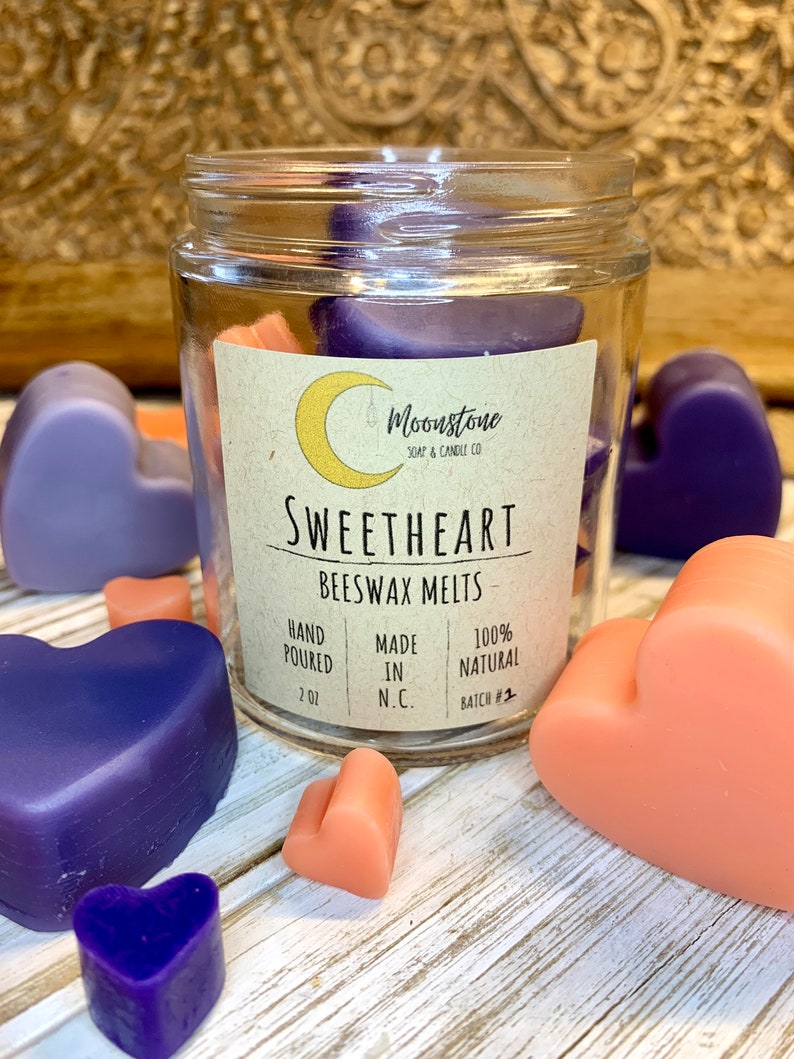 May include: A glass jar filled with heart-shaped beeswax melts in shades of purple and peach. The label reads "Moonstone Soap & Candle Co. Sweetheart Beeswax Melts Hand Poured 2 oz Made in N.C. 100% Natural Batch #2"