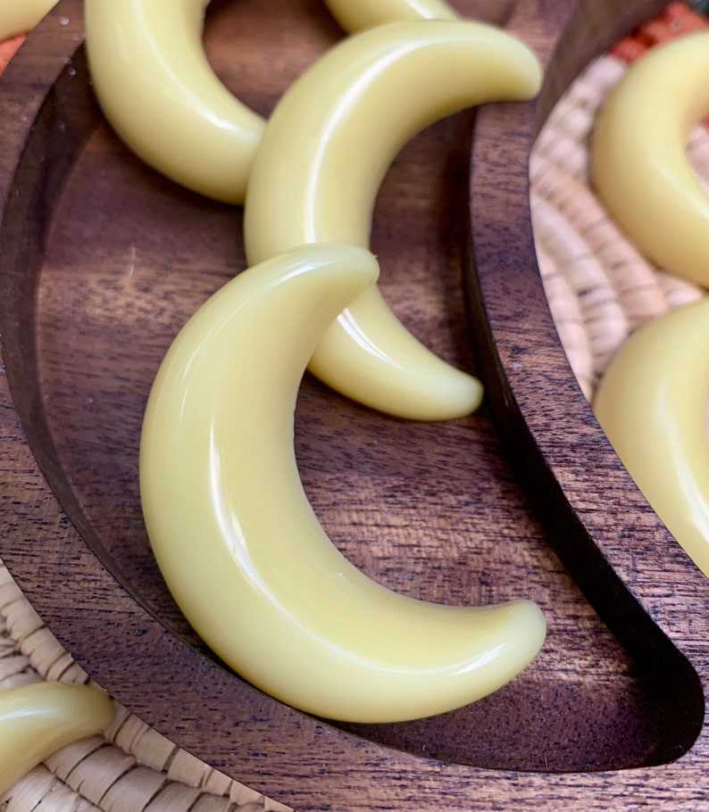 May include: A wooden tray with a crescent moon shape holds multiple small, pale yellow, crescent moon-shaped wax melts.