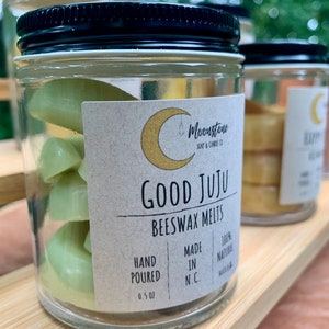 May include: A glass jar with a black lid filled with green beeswax melts. The jar has a label that reads "Moonstone Soap & Candle Co. Good Juju Beeswax Melts Hand Poured 0.5 oz Made in N.C. 100% Natural"