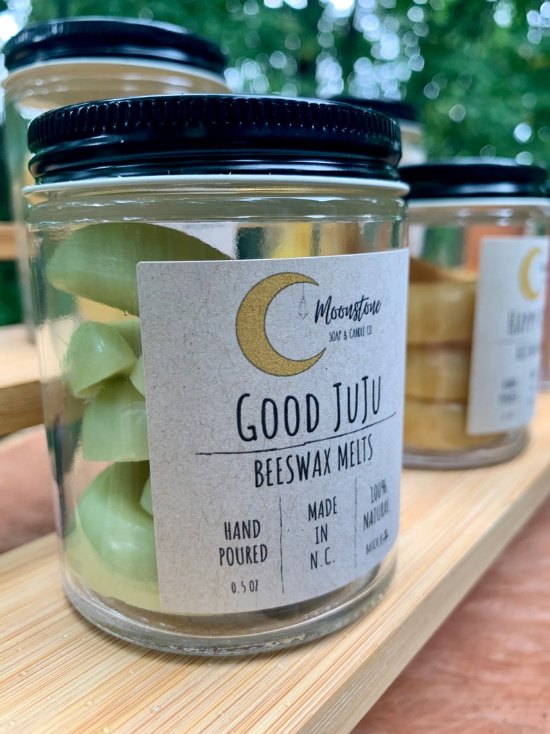 May include: A glass jar with a black lid filled with green beeswax melts. The jar has a label that reads "Moonstone Soap & Candle Co. Good Juju Beeswax Melts Hand Poured 0.5 oz Made in N.C. 100% Natural"