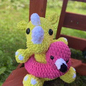 May include: A yellow crocheted rhinoceros holding a pink crocheted flamingo with a white inner tube.