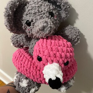 May include: A gray crocheted elephant plush toy sitting on top of a pink crocheted flamingo plush toy. The flamingo has a white beak and black eyes.