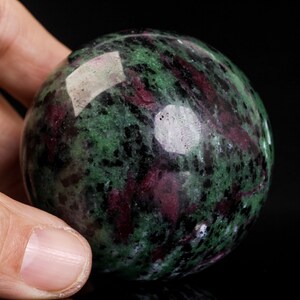 May include: A polished gemstone sphere with a mix of green, dark red, and black colors. The sphere is held in a hand, and the surface reflects light. The stone appears to be a type of ruby.