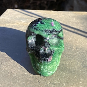 May include: Green and black gemstone skull with pink accents. The skull is carved from a natural stone and has a detailed design.