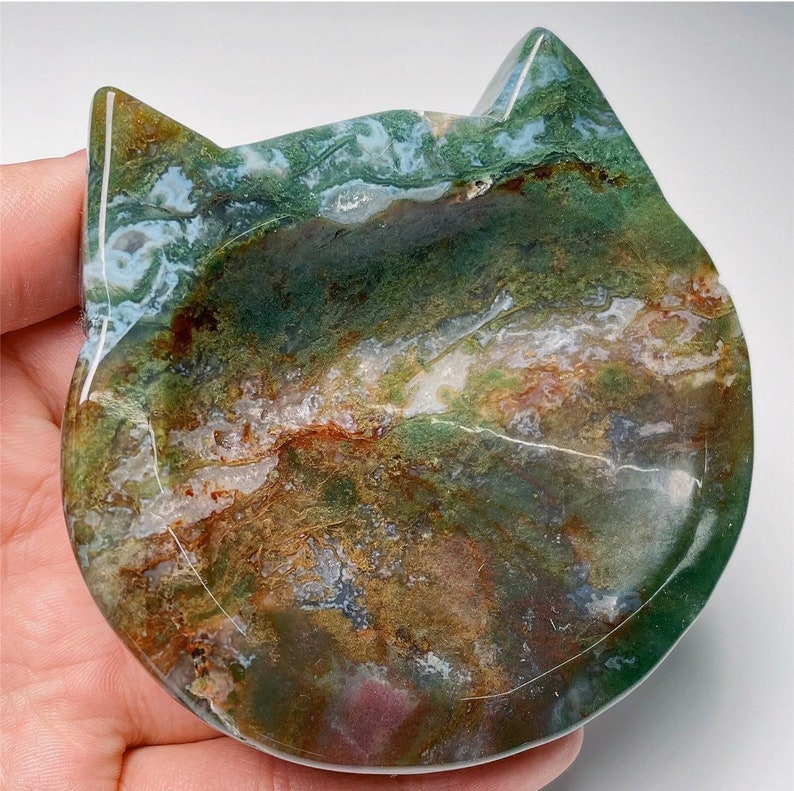 May include: A green, brown and white agate cat dish. The dish is carved in the shape of a cat's head and has a smooth, polished surface.