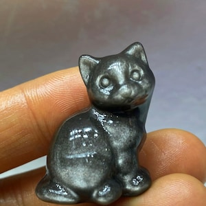 Puede incluir: Una pequeña figurita de gato negro y brillante hecha de obsidiana. El gato está sentado con la cabeza ligeramente inclinada hacia un lado.
