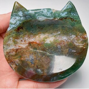 May include: A green, brown and white agate cat dish with a smooth, polished surface. The dish is shaped like a cat's face and has a shallow, rounded interior.