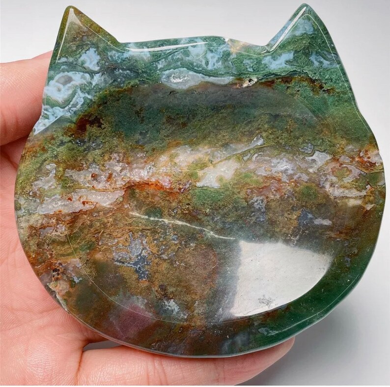 May include: A green, brown and white agate cat dish with a smooth, polished surface. The dish is shaped like a cat's face and has a shallow, rounded interior.