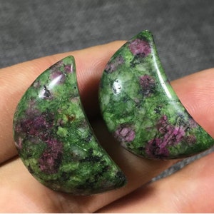 Może przedstawiać: Dwa polerowane kamienie szlachetne w kształcie półksiężyca. Kamienie mają mieszankę zielonych i głębokich czerwonych odcieni, z marmurkowym wyglądem. Gładkie, zakrzywione powierzchnie odbijają światło, podkreślając naturalne wzory. Prawdopodobnie używane do biżuterii lub celów dekoracyjnych.