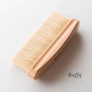 May include: A wooden brush with natural bristles. The brush has a curved wooden handle with the word "Buffy" engraved on it.