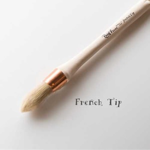 May include: A wooden-handled paint brush with a copper ferrule and a natural bristle brush head. The brush is labeled "French Tip" and "Real Fine" in cursive writing.