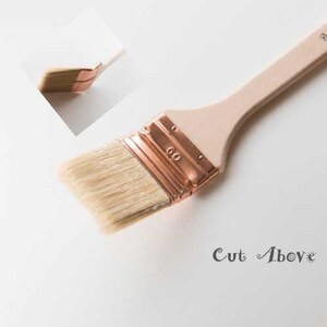 May include: A paint brush with a light wood handle and natural bristles. The brush head is secured with a copper-colored metal band. The text "Cut Above" is printed in a script font near the brush.