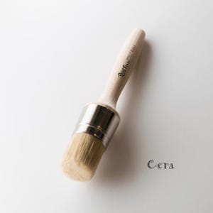 May include: A close-up of a round, white, natural bristle paint brush with a silver metal ferrule and a wooden handle. The handle is labeled "Real Milk Paint Co."