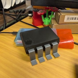 May include: A black electronic component with four silver-colored legs is the focal point. The component sits on a wooden surface, with other objects like a red box and a 3D-printed flower nearby. The background includes a computer monitor.