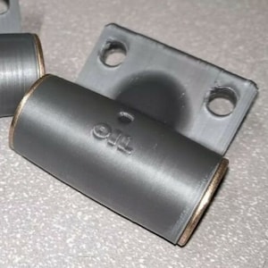 May include: A gray metal pipe fitting with a gold-colored edge and the word "The" printed on the side. The fitting is attached to a gray metal plate with two holes.
