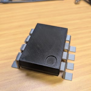 May include: A black rectangular electronic component with a circular indentation on top. It is supported by grey, rectangular pins. This component is likely used in electronics or circuit boards.