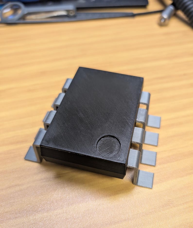 May include: A black rectangular electronic component with a circular indentation on top. It is supported by grey, rectangular pins. This component is likely used in electronics or circuit boards.