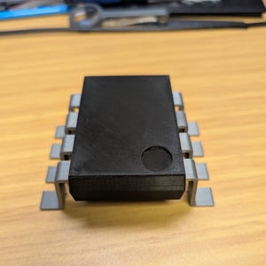 May include: A black electronic component with a rectangular body and grey metal pins. The component has a circular indentation on its top surface. The item is placed on a light brown wooden surface.